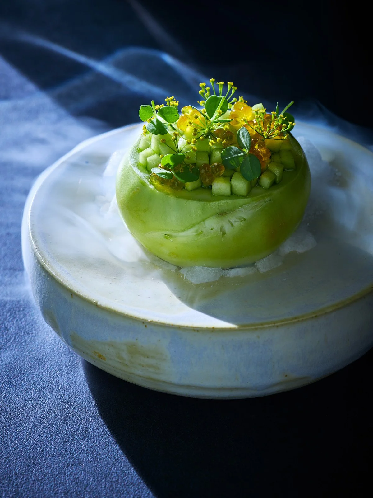A green apple with topping of diced cucumber, yellow edible flowers, green leaves, and small yellow berries, served on a white plate with ice.