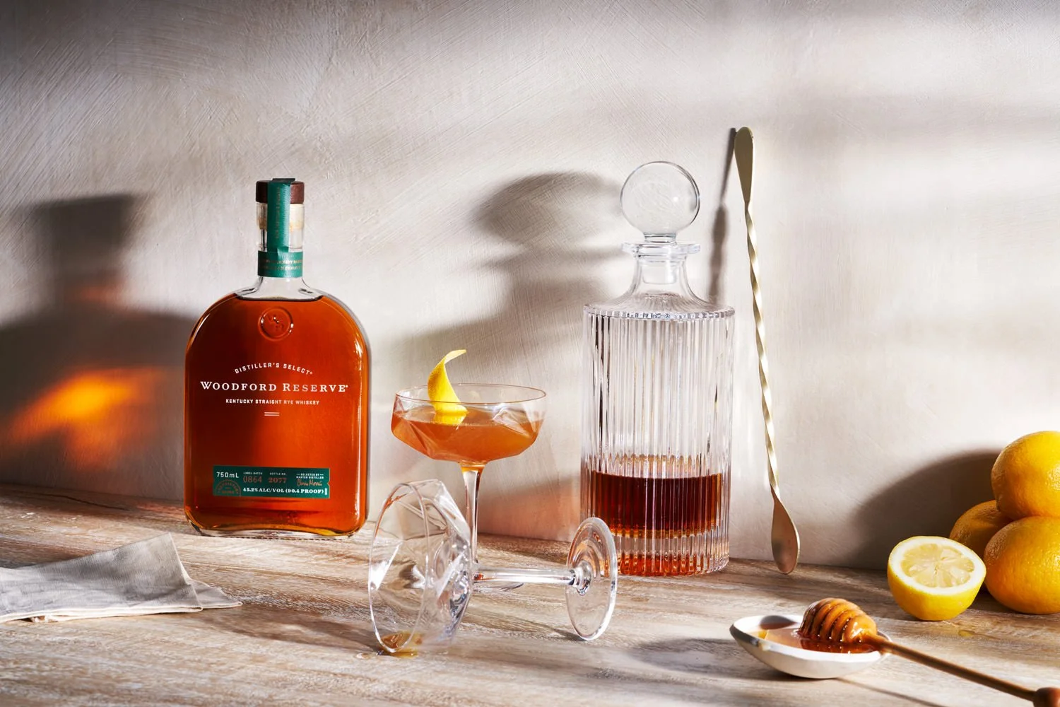 A still life with a bottle of Woodford Reserve bourbon, a cocktail glass with lemon peel, a glass decanter with bourbon, a silver stirring spoon, lemons, and honey on a wooden surface and a light-colored wall background.