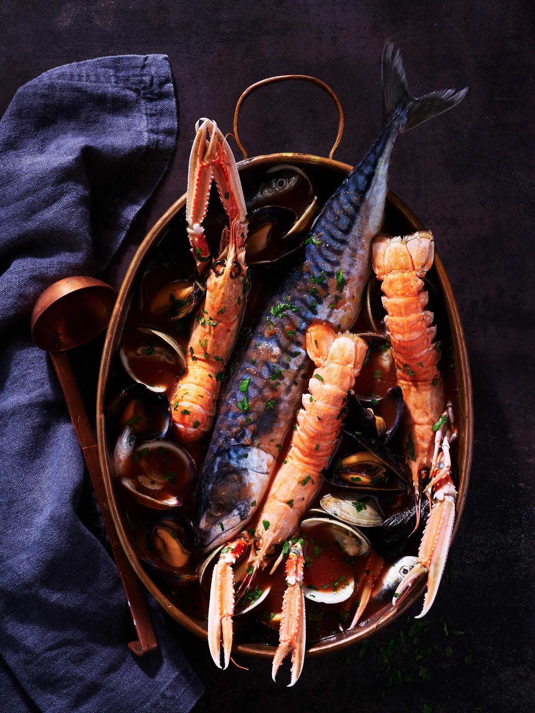 A seafood dish with a whole fish, lobster, shrimp, and clams in a dark bowl garnished with herbs.