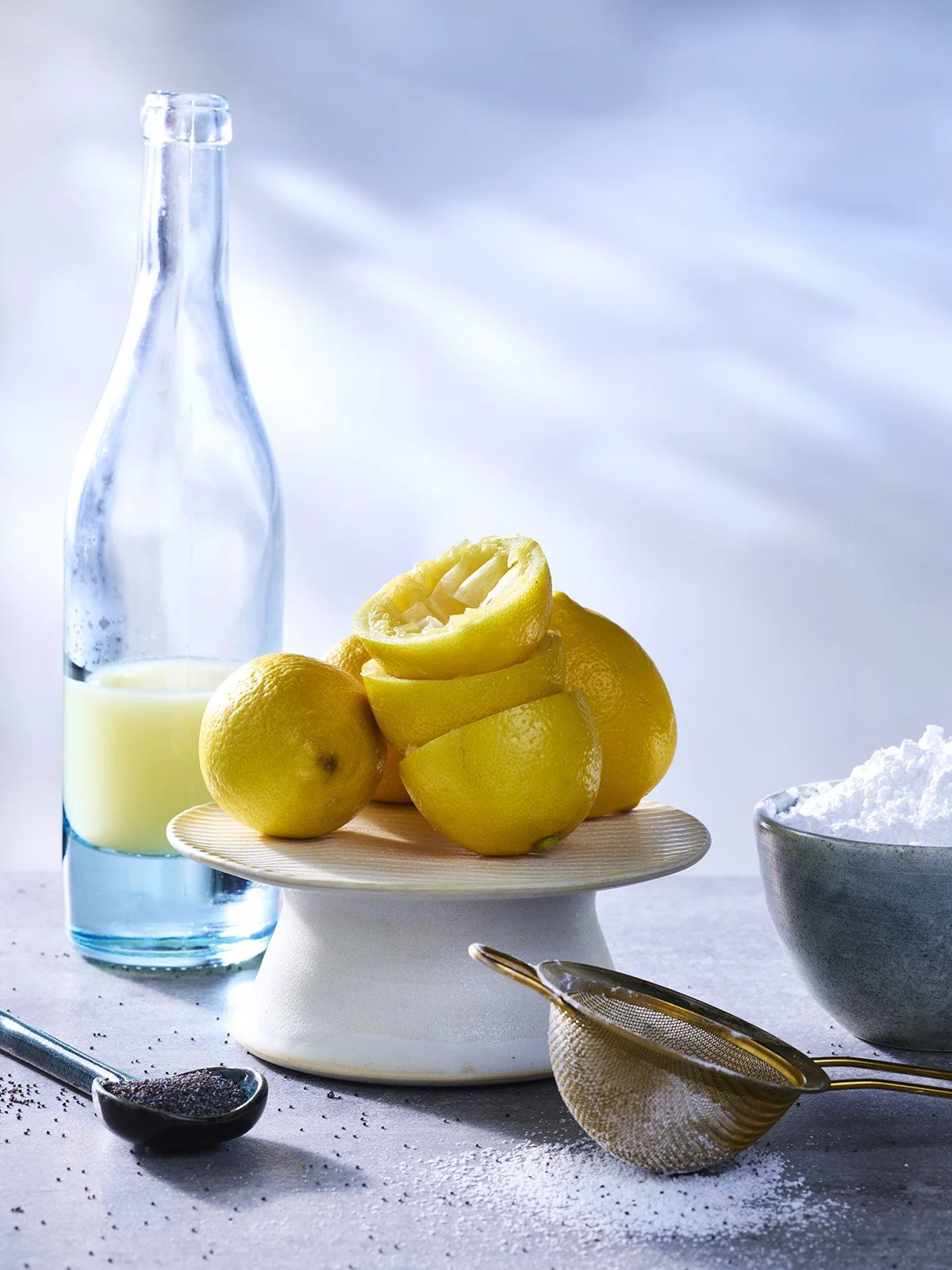 A still life composition featuring a glass bottle of lemon juice, sliced lemons on a pedestal, a bowl of powdered sugar, a spoon with poppy seeds, and a small sieve, all set on a light surface with a soft, cloudy sky background.