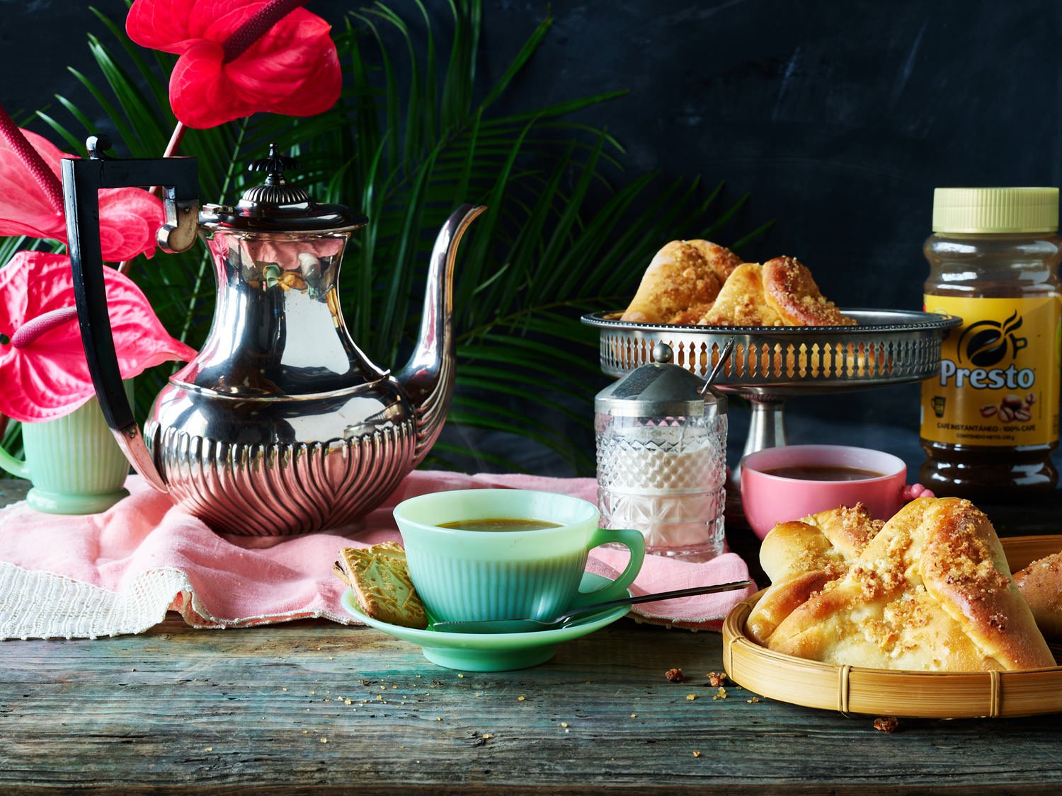 A tea setting a wooden table with a pink cloth, with bread or pastries on a silver stand in the background.