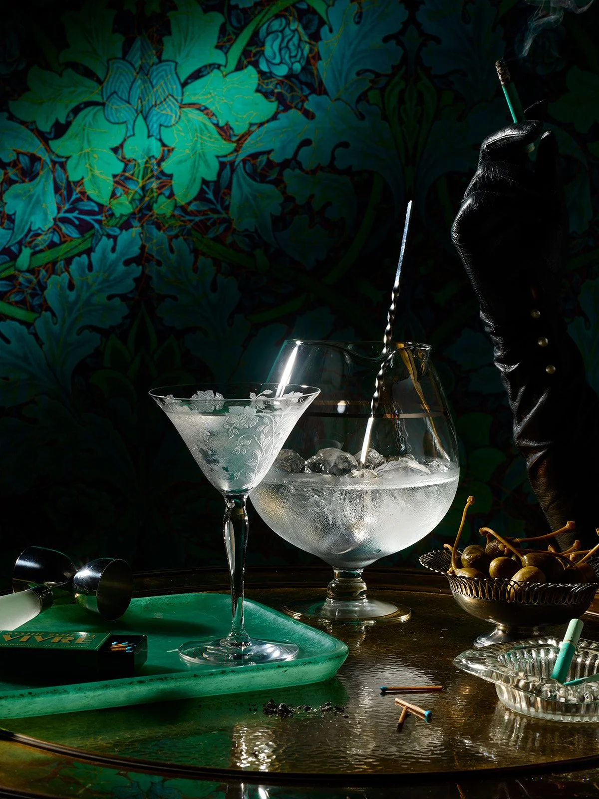 A hand wearing a black leather glove is holding a lit cigarette over a large glass bowl of ice water. There is a smaller martini glass, a green tray with bar tools, and a bowl of cherries on a reflective table, with a dark floral wallpaper in the bac