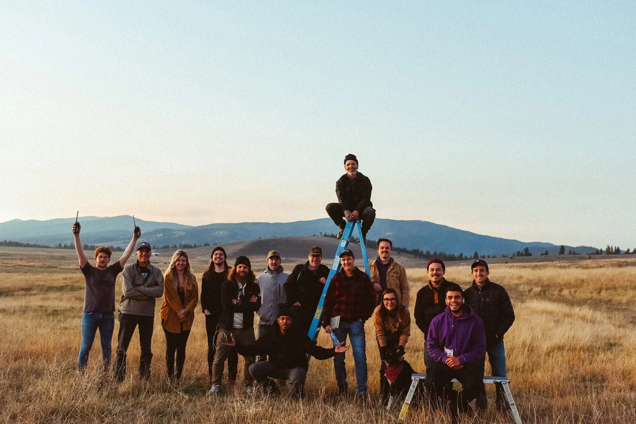 Group of people outdoors on a grassy field with mountains in the background. One person is sitting on top of a ladder while others pose around and under it. Some are raising their arms or holding objects.