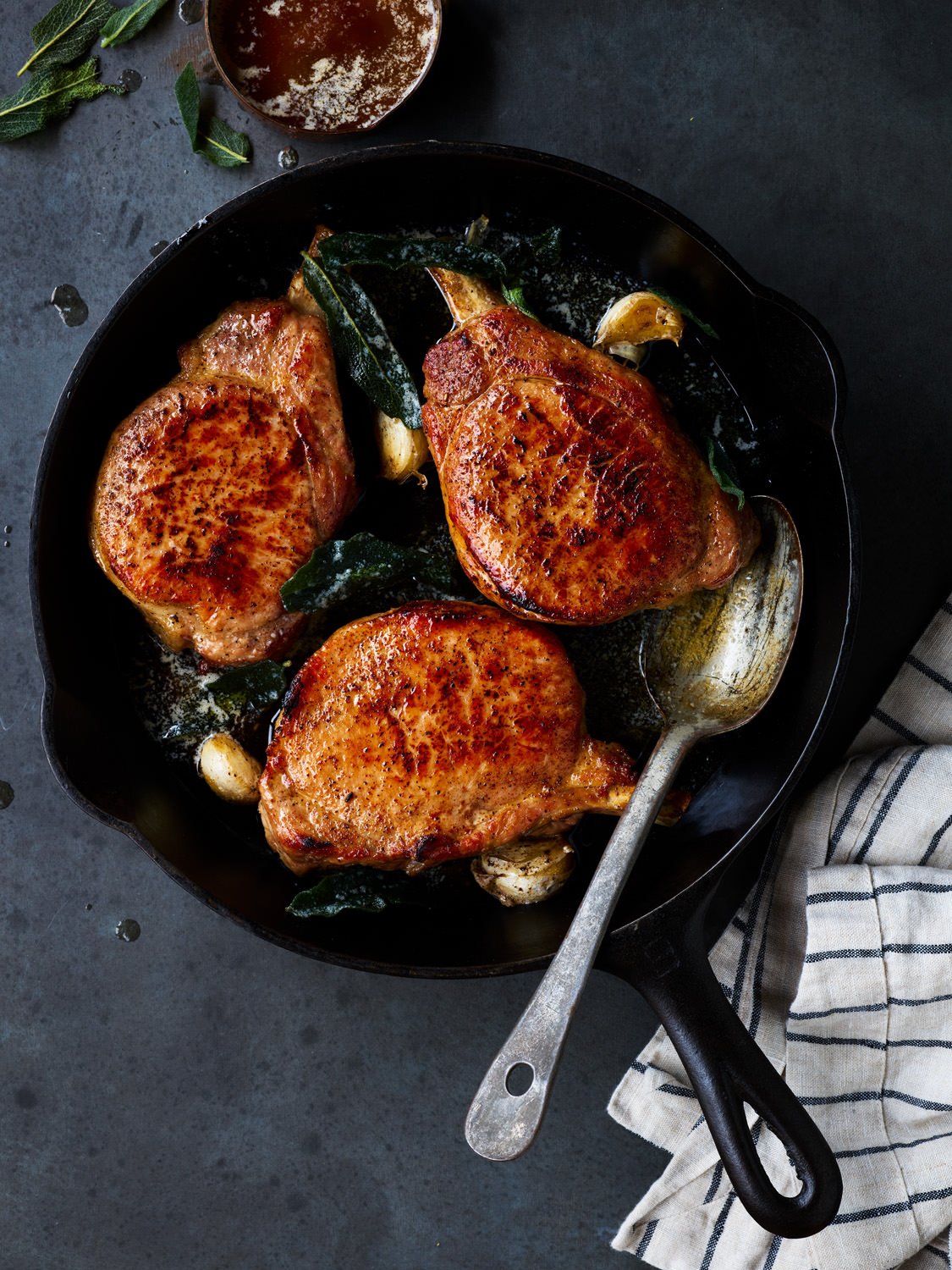 Pan with three cooked chicken breasts, garlic cloves, and sage leaves, on a dark surface with a striped cloth and a small bowl of seasoning.