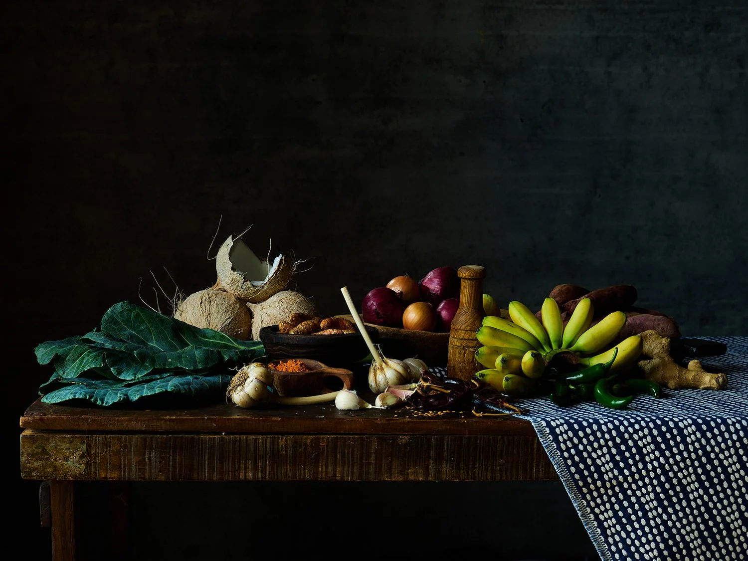 Arrangement of fresh vegetables and fruits on a dark wooden table with a blue and white tablecloth, including bananas, garlic, onions, sweet potatoes, ginger, green peppers, and leafy greens.
