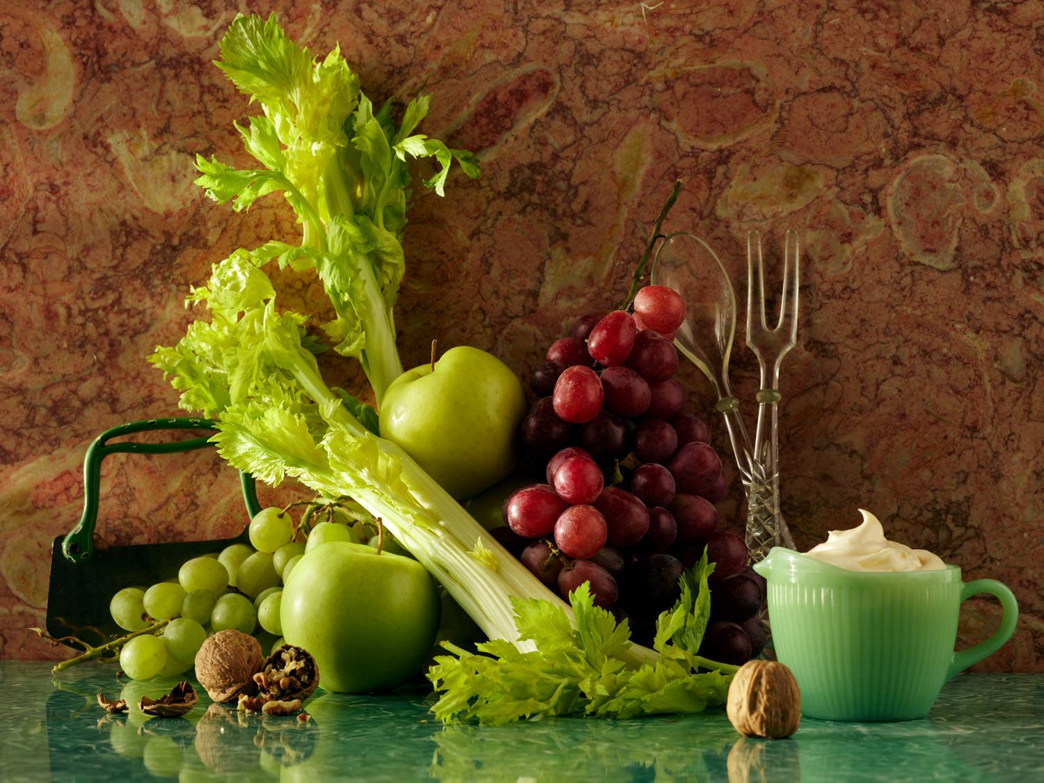 A still life arrangement of green apples, a bunch of red grapes, celery, walnuts, and a green cup filled with cream, set on a green marble surface with a pink marbled background.