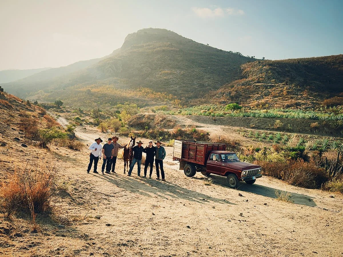 Group of five men, one with a horse, standing in a dry, mountainous landscape with a red pickup truck nearby, under a clear sky.