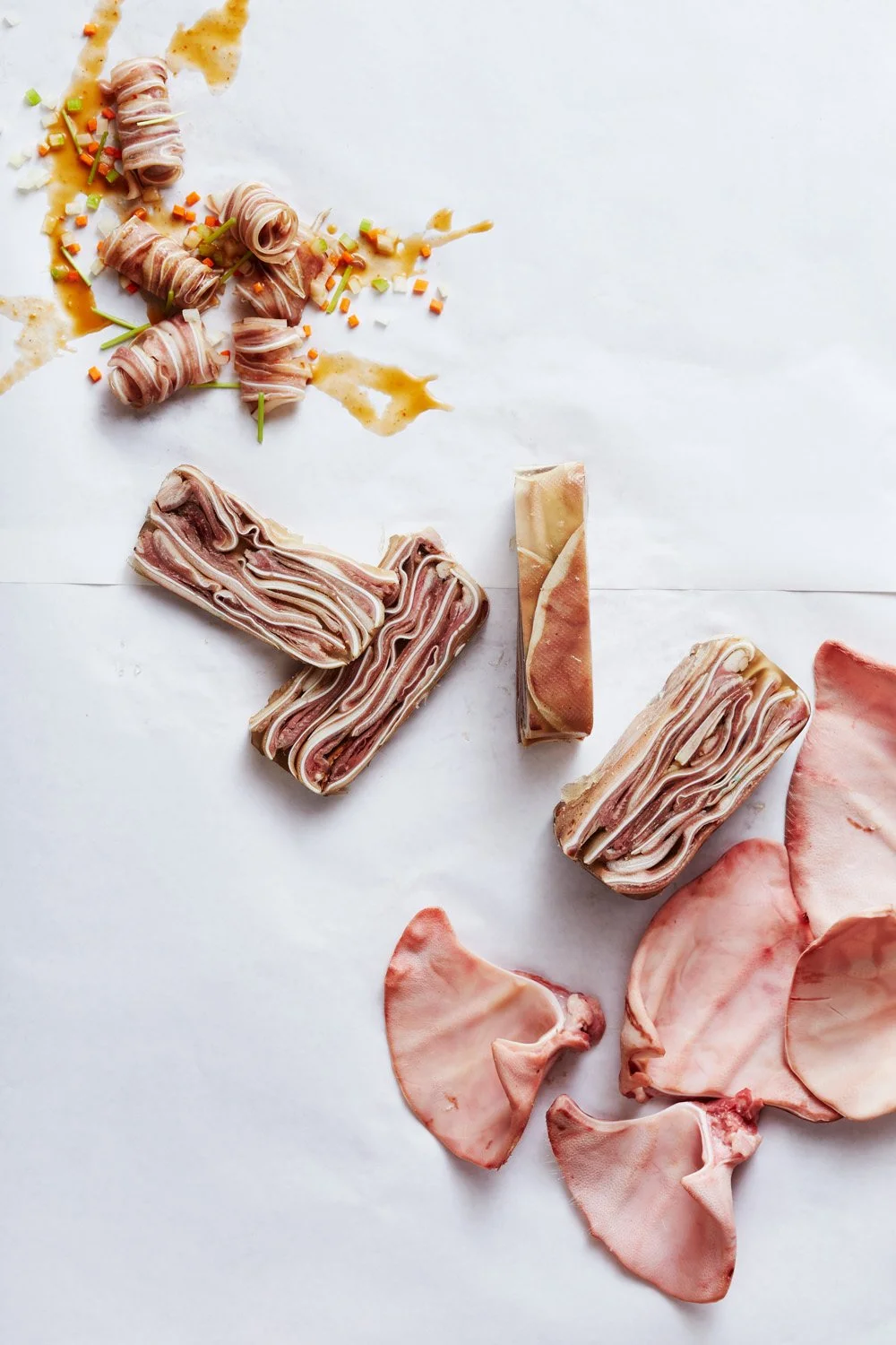 Assorted sliced meats, including bacon and ham, arranged on white paper with some pieces rolled or folded, some stainless steel sauce or gravy, and colorful chopped vegetables.