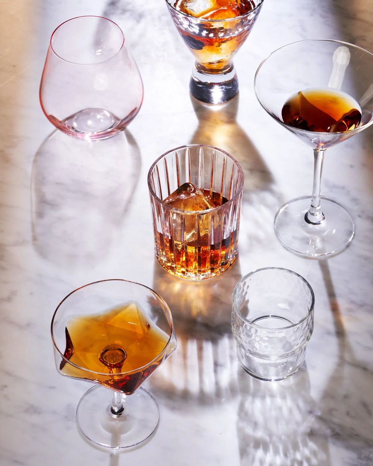 Seven different glasses and a tumbler with ice and amber-colored beverages on a white marble surface.