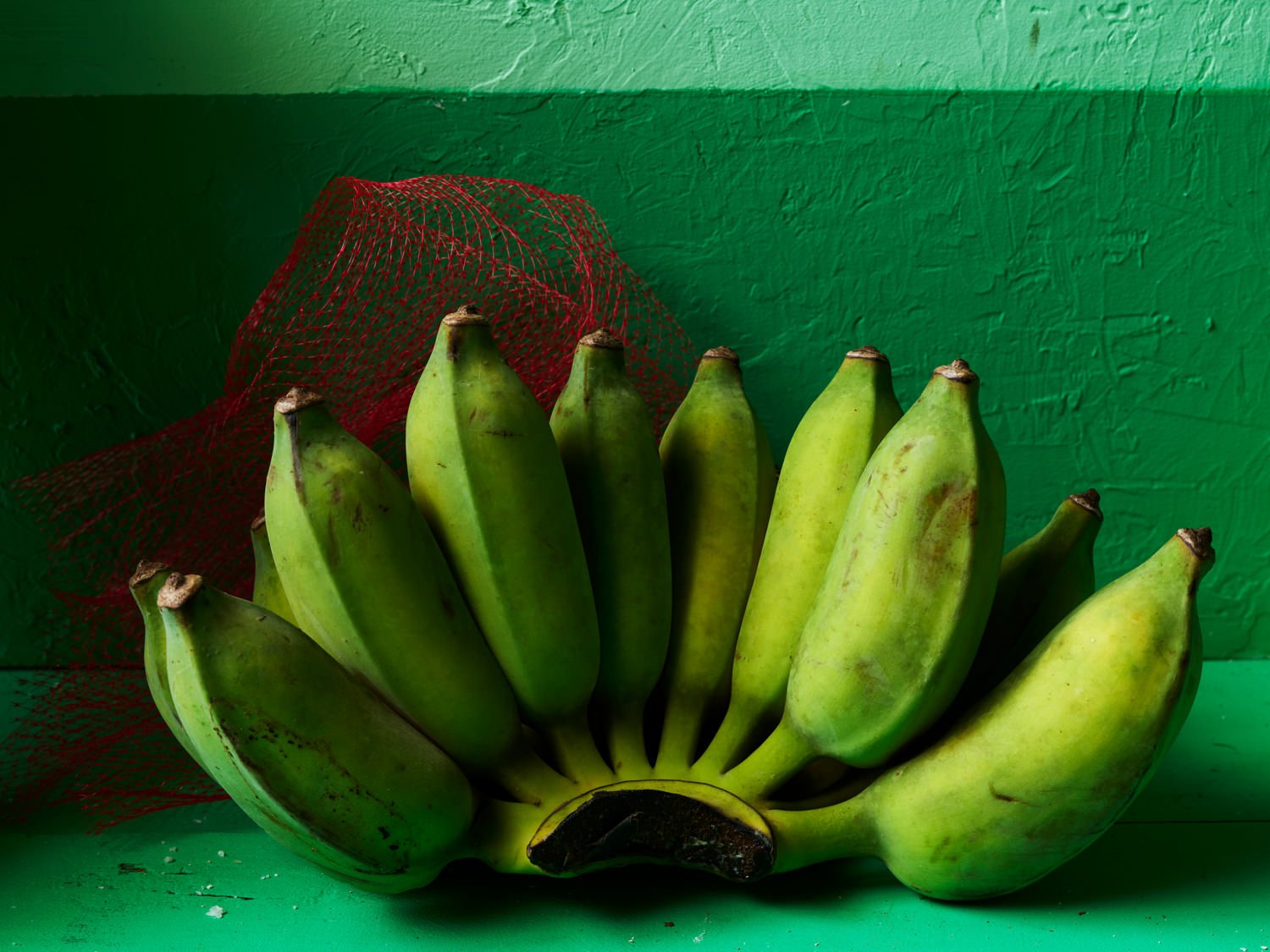 A bunch of green bananas resting on a green surface against a green wall, with a red net in the background.