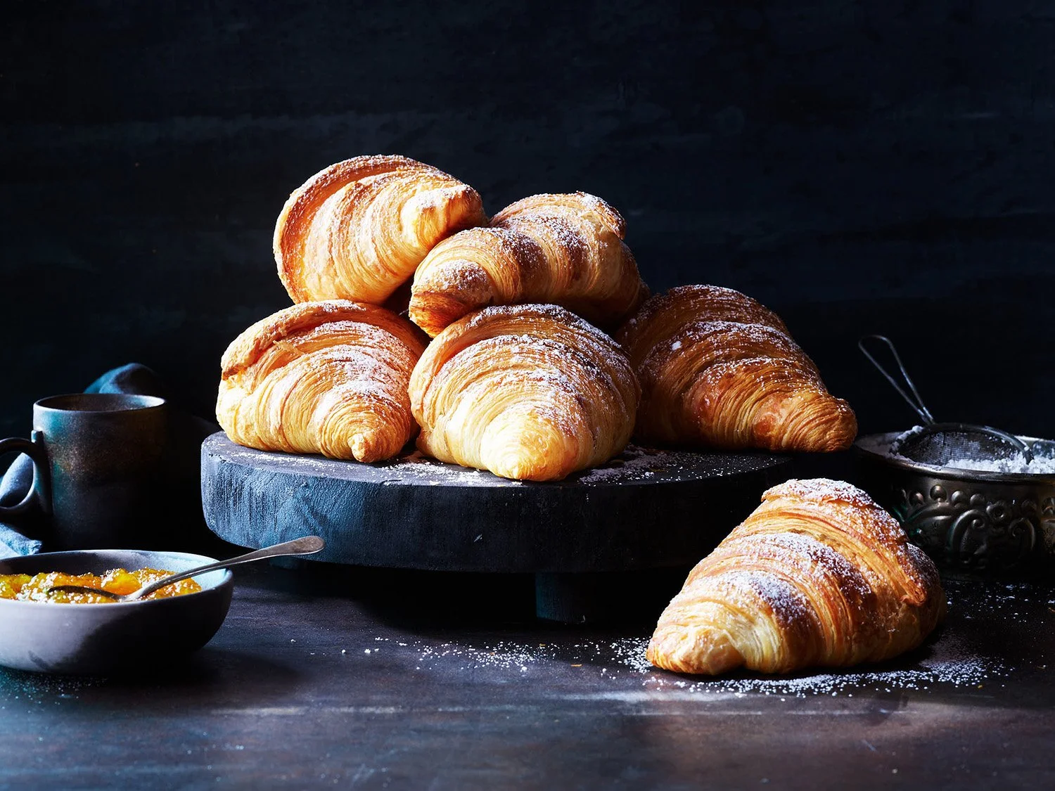 Fresh croissants stacked on a black round board with powdered sugar, with a dark background and a bowl of fruit jam and a coffee mug nearby.