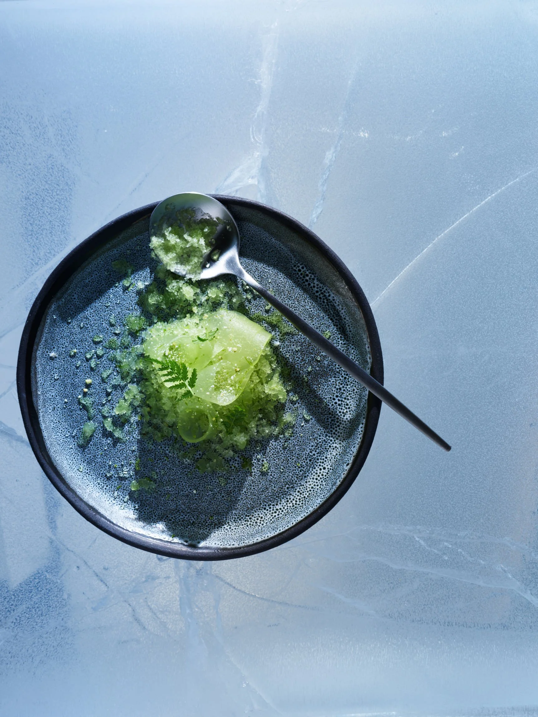 A black bowl filled with green vegetables and herbs, with a metal spoon resting on the edge, on a light blue surface with a frosted pattern.