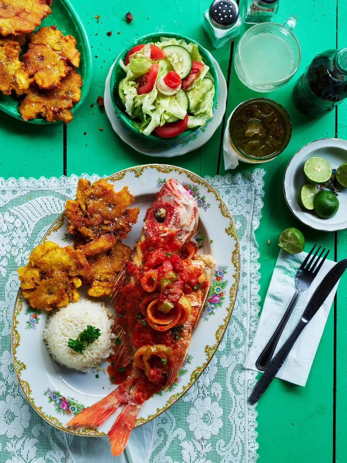 A plate of cooked fish topped with tomato and bell pepper sauce, rice garnished with a sprig of parsley, and fried plantains. A side salad of lettuce, cucumber, and tomato, along with lime wedges, is on the side. Beverages including sodas and a glass