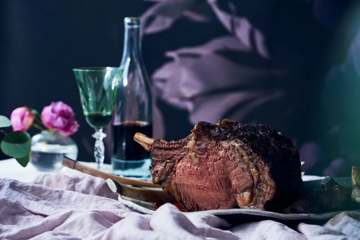 A roasted prime rib sitting on a plate with a carving fork, on a table with a pink cloth, glasses of wine, a vase with pink flowers, and a dark background.