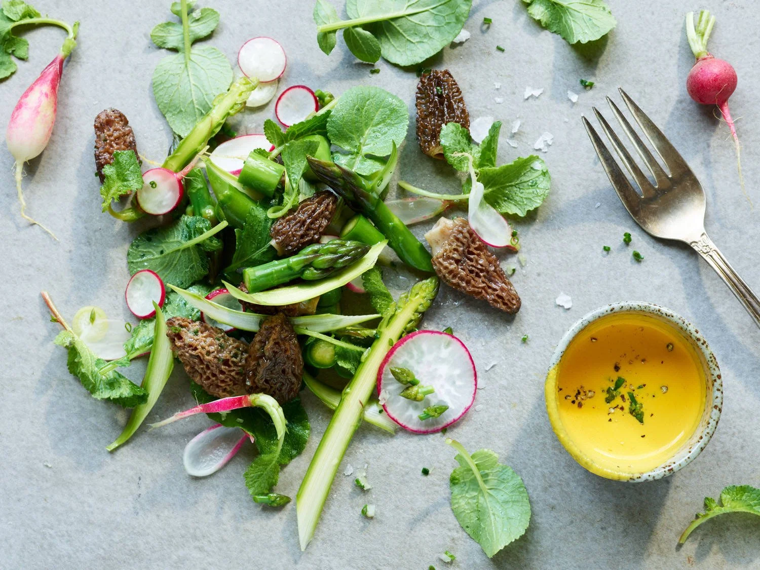 Fresh vegetable salad with radishes, asparagus, lettuce, and edible morel mushrooms on a gray surface, with a small bowl of yellow vinaigrette and a fork nearby.