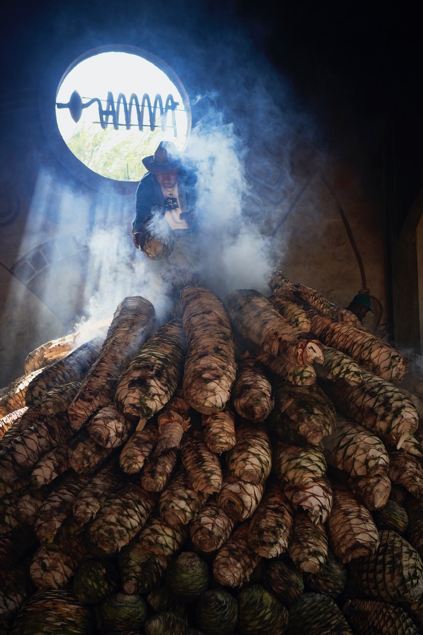 A person wearing a hat and jacket standing among a large stack of smoked, cylindrical-colored yams or sweet potatoes with smoke rising around them, in front of a bright circular window with a lightning bolt symbol.