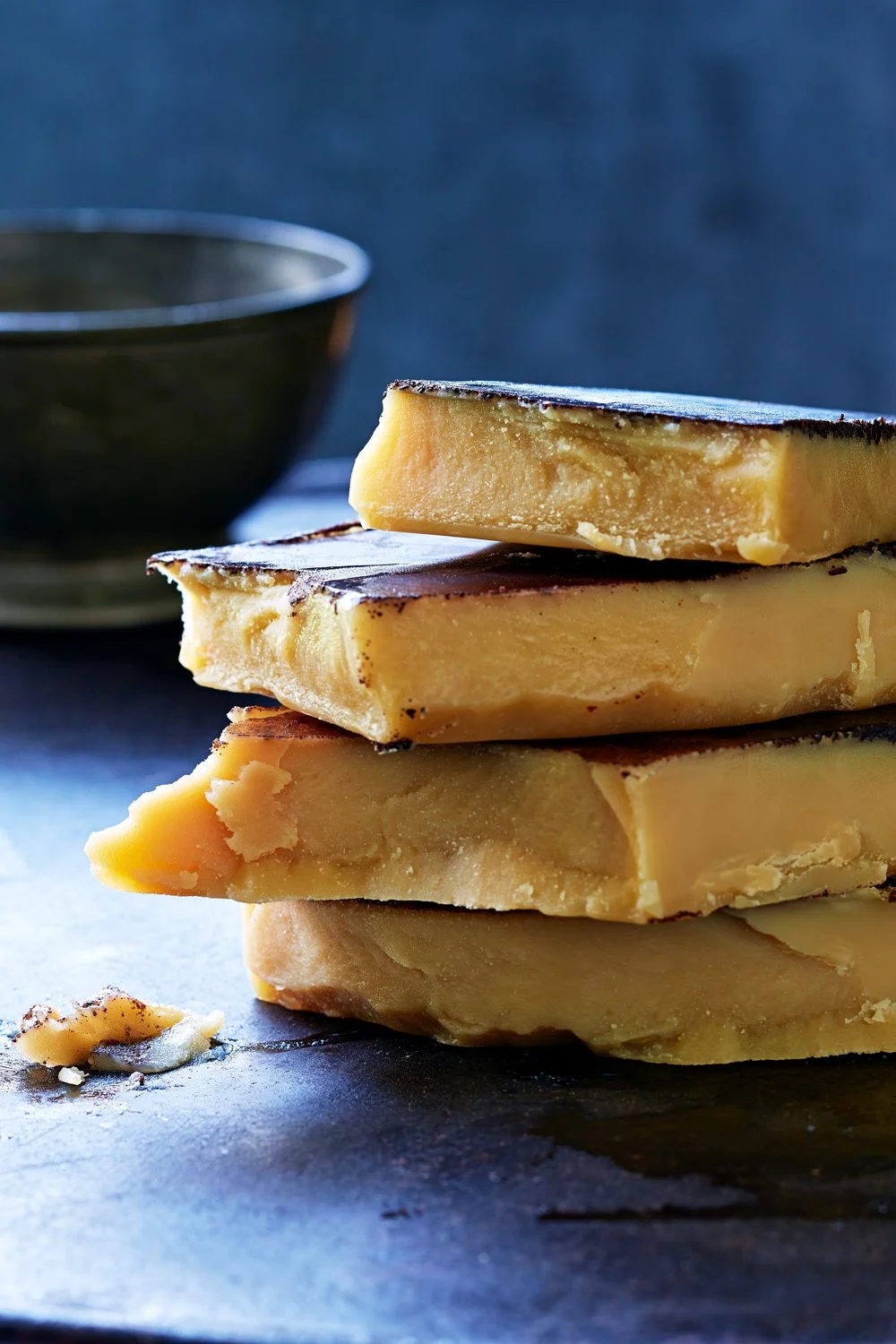 Stacked pieces of white to yellow cheese with a dark bowl in the background.
