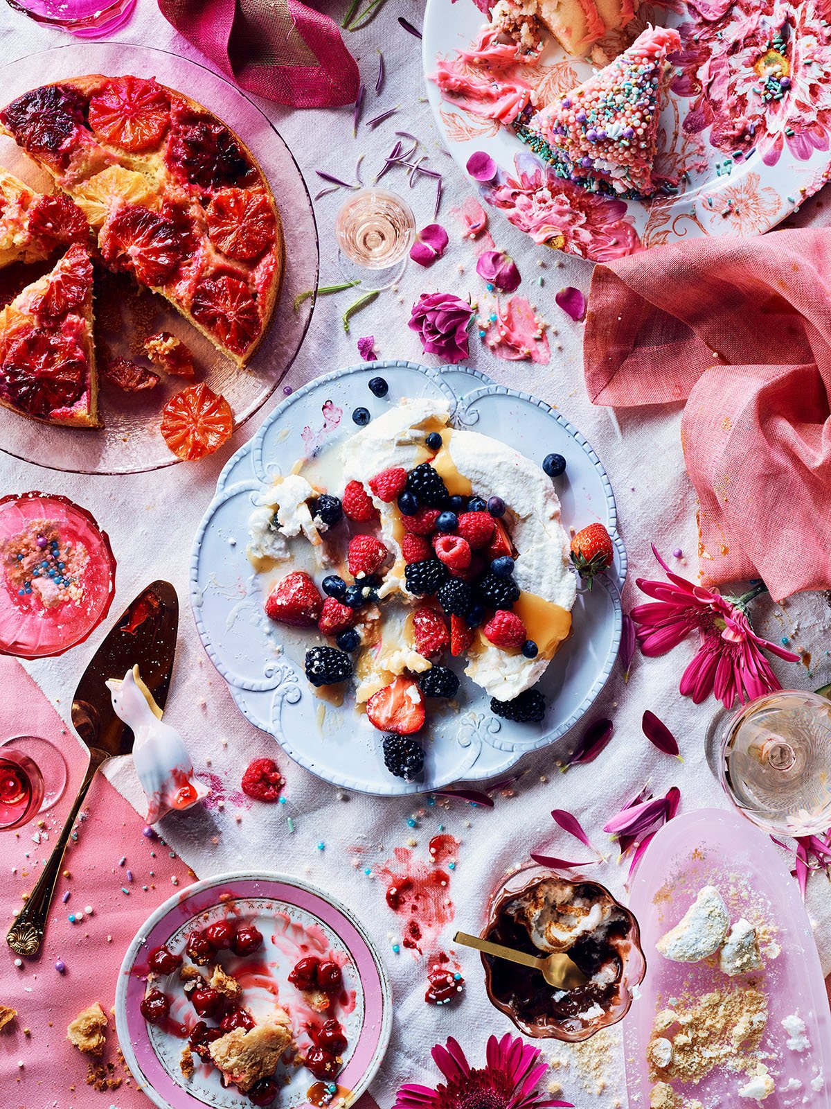 Partially eaten berry dessert, pizza slice with caramel and citrus slices on a glass plate, decorated with flowers and sprinkles, scattered crumbs and dessert remnants on the table.