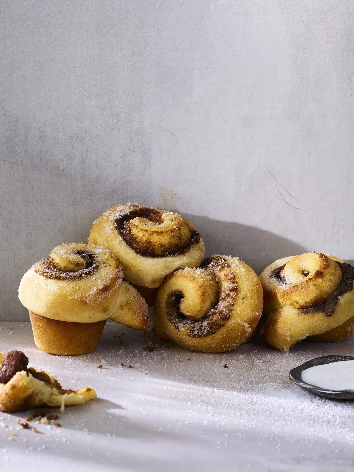 Cinnamon rolls with icing sugar on a white sheet, some broken pieces, with a spoon of sugar.