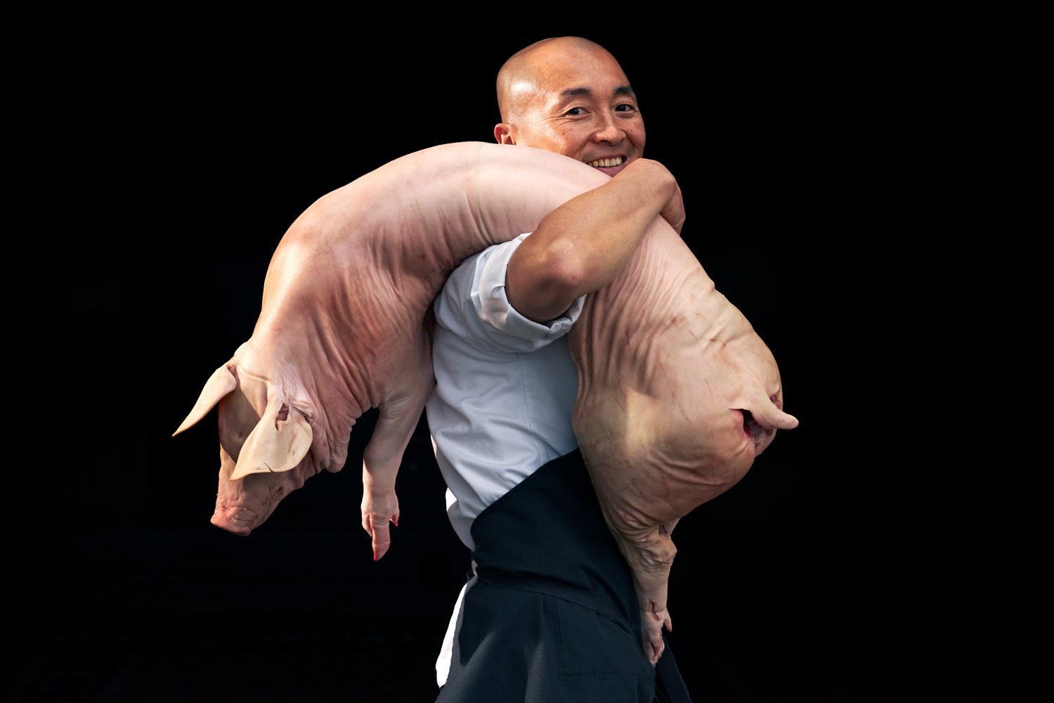 A man smiling while carrying a large pig on his shoulder against a black background.