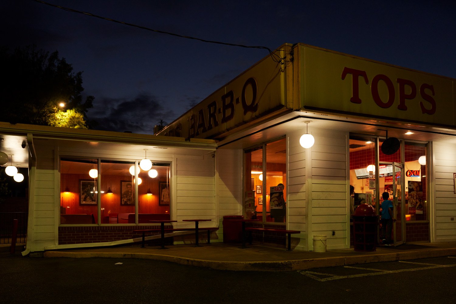 Night scene of a fast-food restaurant with illuminated interior and exterior lights, with visible signs that read 'TOPS' and 'BARBQ'. The restaurant has white wooden siding and large front windows through which people inside can be seen.
