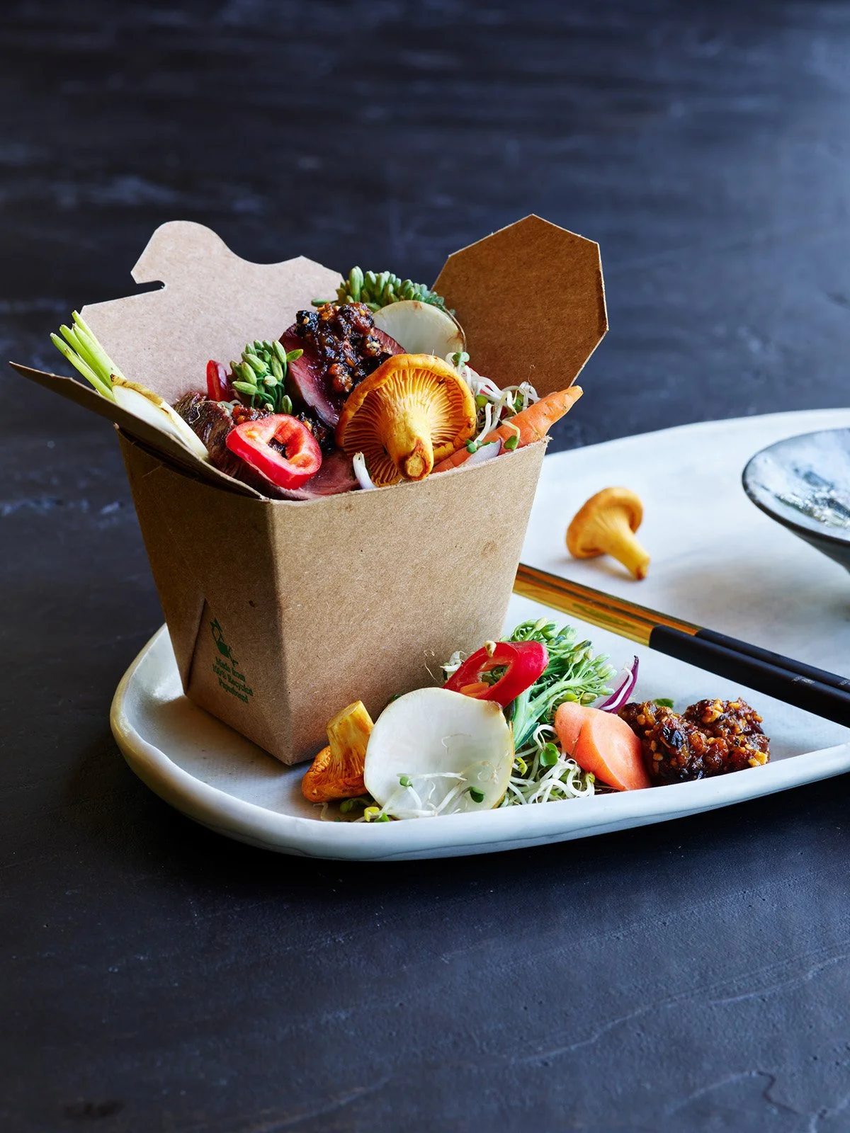 A takeout container filled with Asian-inspired ingredients, including mushrooms, vegetables, and microgreens, placed on a white plate with chopsticks and a side of vegetables.