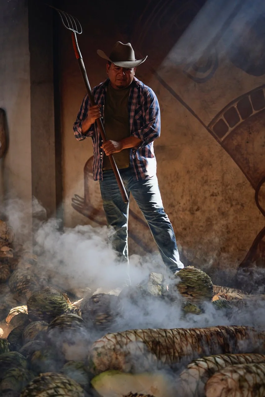 A man dressed as a farmer, wearing a cowboy hat, plaid shirt, and jeans, holding a pitchfork, standing amidst steaming vegetables or produce in a rustic setting with a mural background.