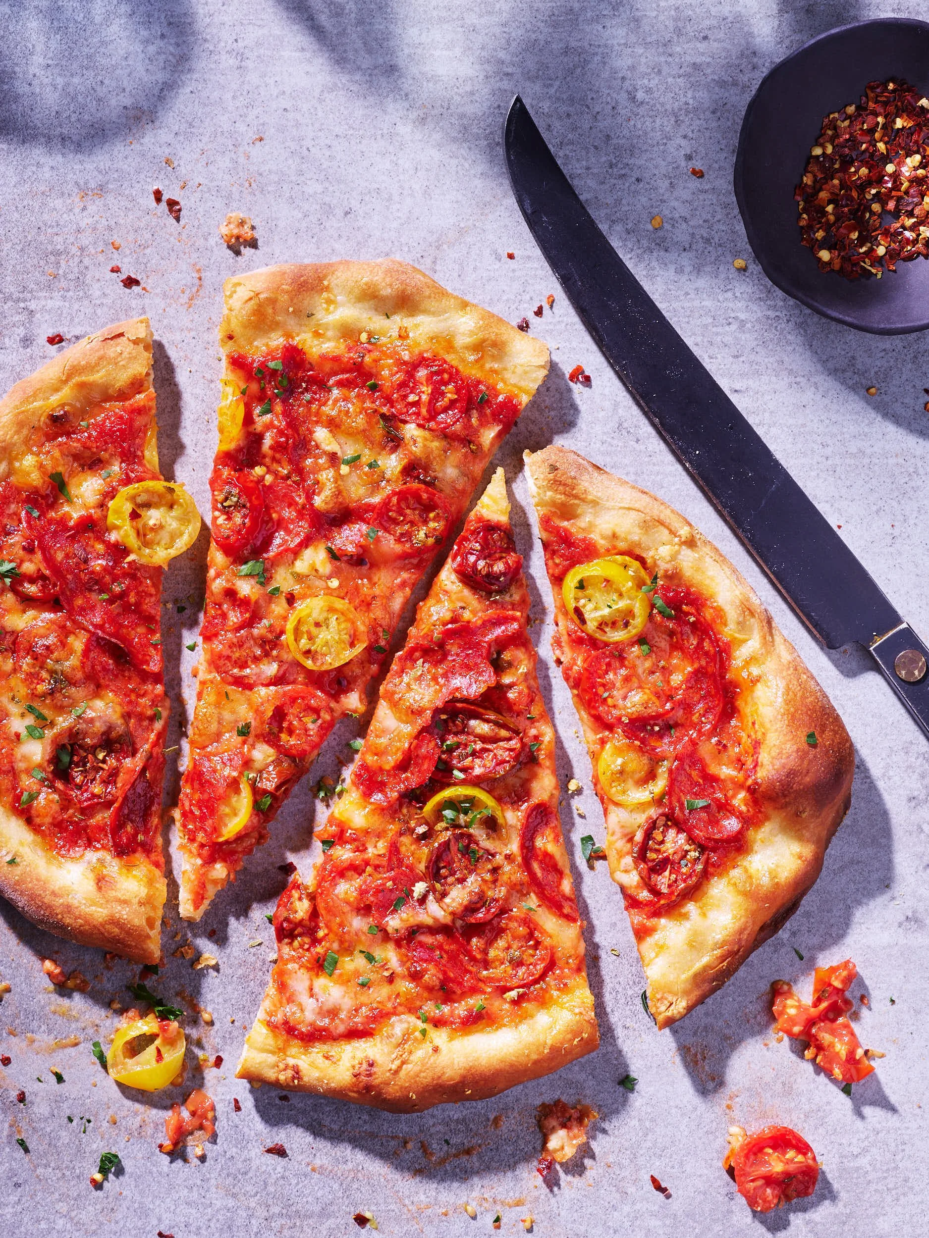 A partially eaten pepperoni and yellow pepper pizza on a gray surface, with a black knife and a small black bowl of crushed red pepper flakes.