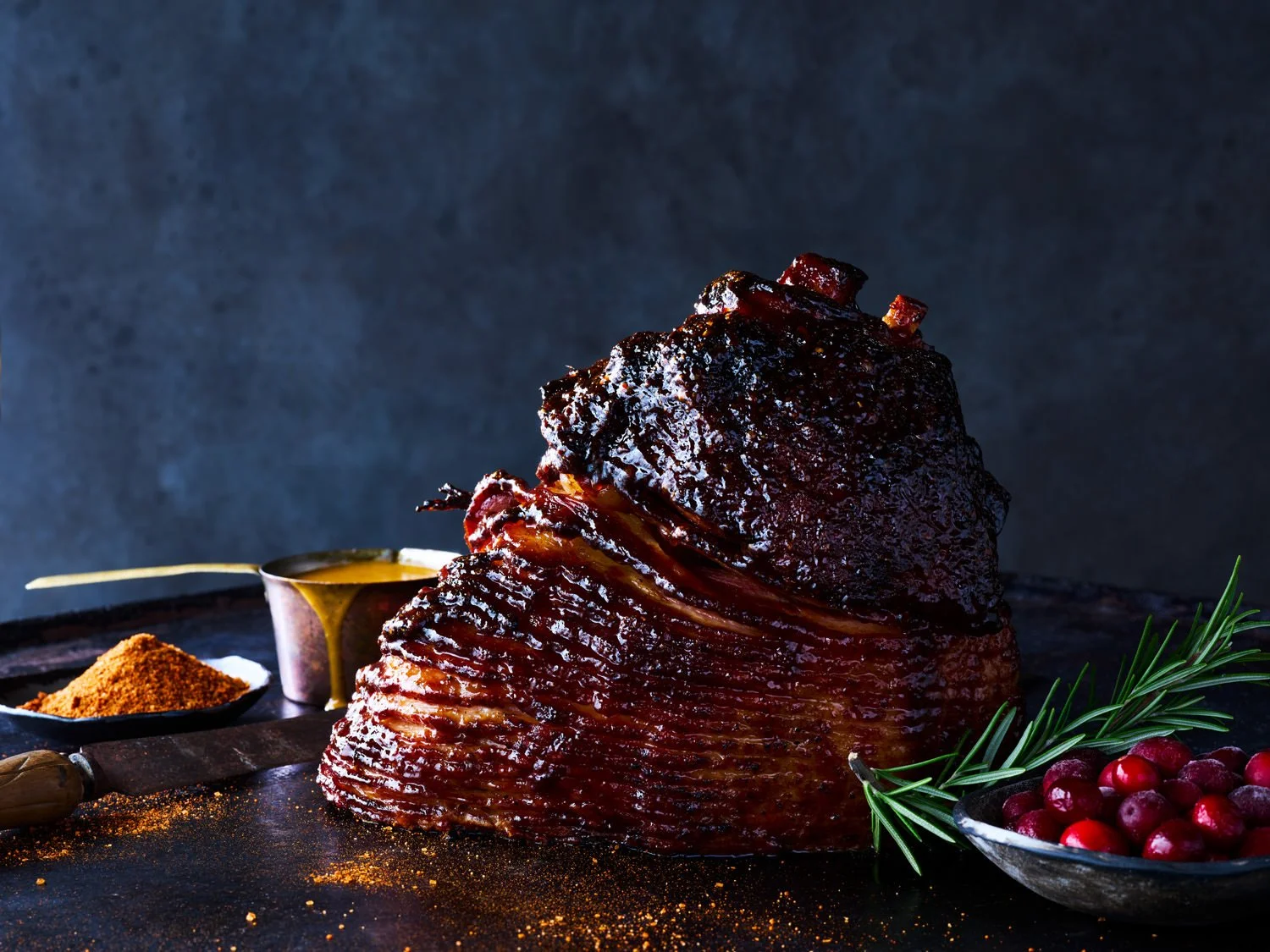 A large glazed smoked beef brisket on a dark surface with sprigs of rosemary, red berries, a small bowl of cranberry sauce, a ramekin of mustard, and spices.