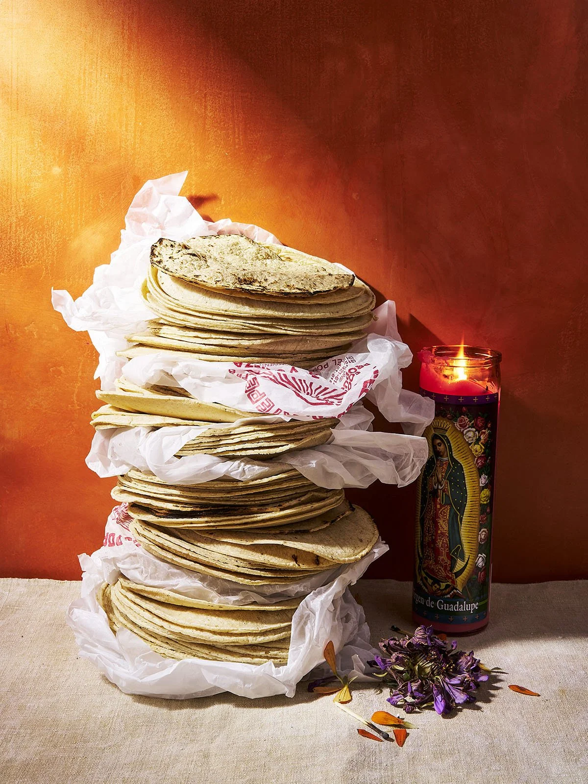 A stack of corn tortillas on crumpled white paper, next to a lit candle with an image of Our Lady of Guadalupe, on a beige cloth table with purple and orange flowers nearby, against an orange wall.