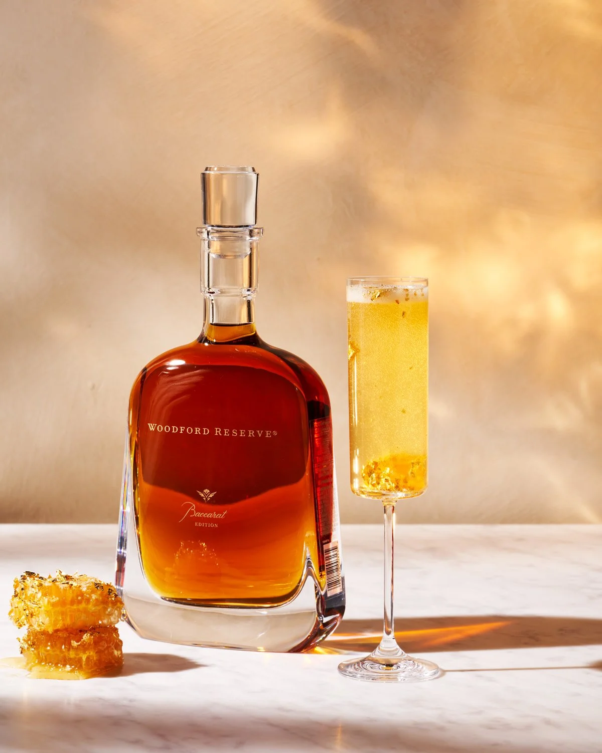 A bottle of Woodford Reserve bourbon whiskey, a glass of champagne, and honeycomb on a white surface with a gold-toned background.
