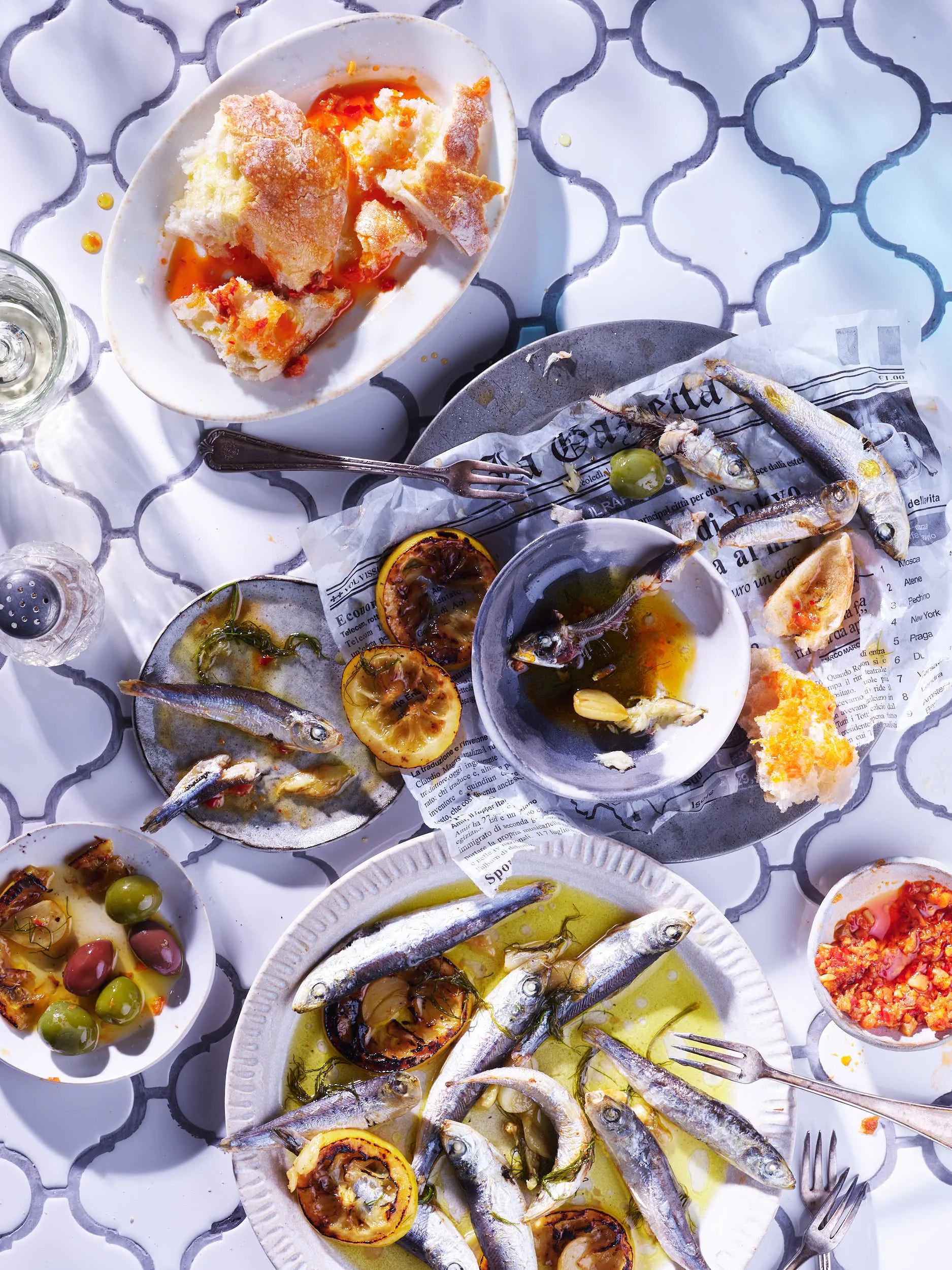 Seafood meal with small fish, lemon slices, olives, and what appears to be bruschetta with tomato topping served on various plates and bowls on a patterned table.