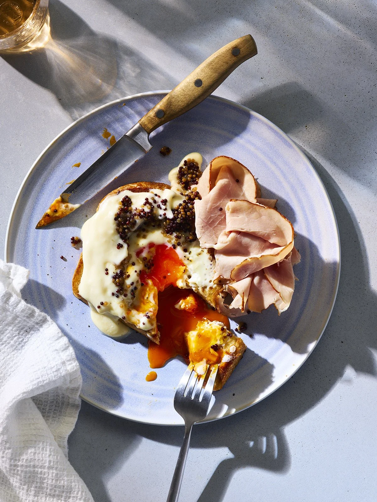 A plate with a breakfast dish featuring a fried egg, sliced ham, and a piece of toast topped with melted cheese and mustard seeds, with a fork and knife, on a light-colored surface.