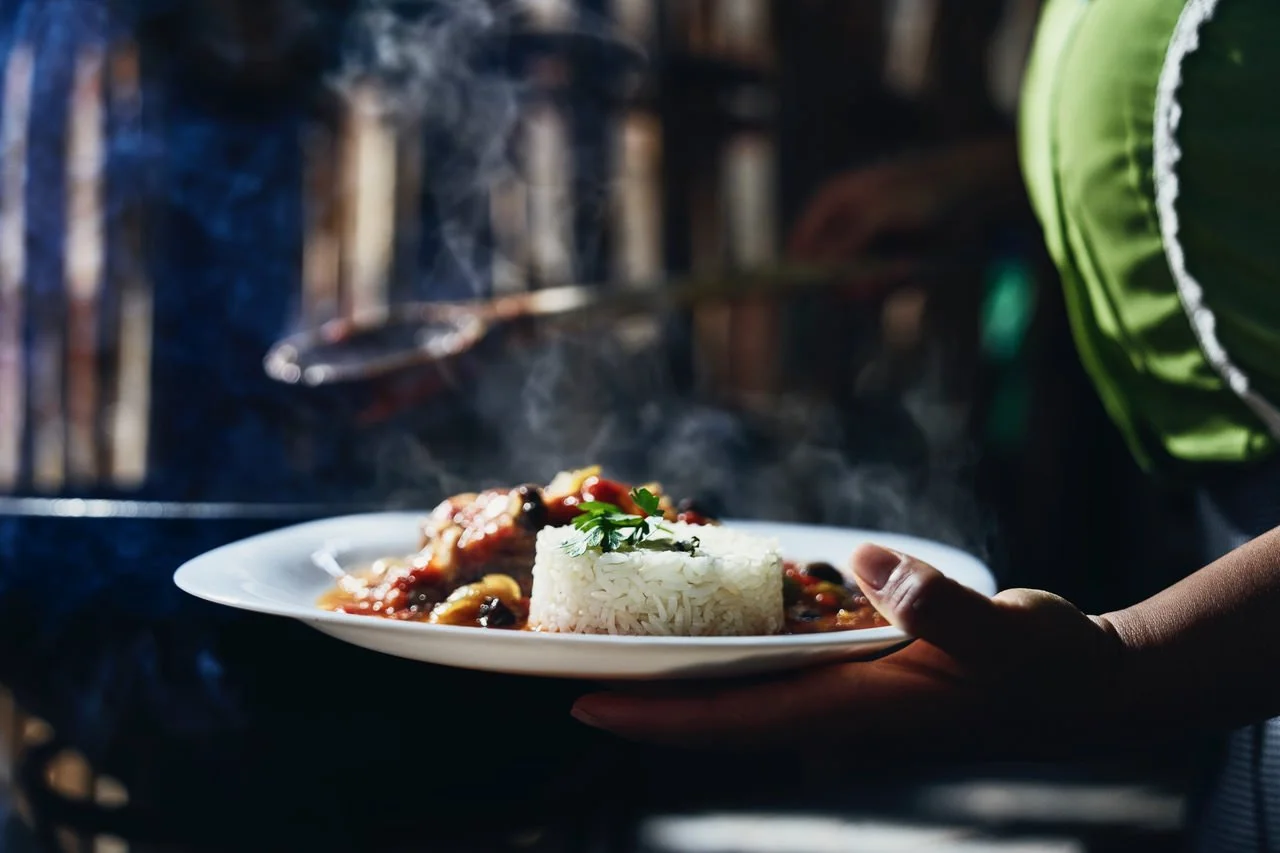 A person holding a plate of steaming rice with a vegetable and meat stew garnished with a sprig of cilantro in front of a smoky, outdoor kitchen setting.