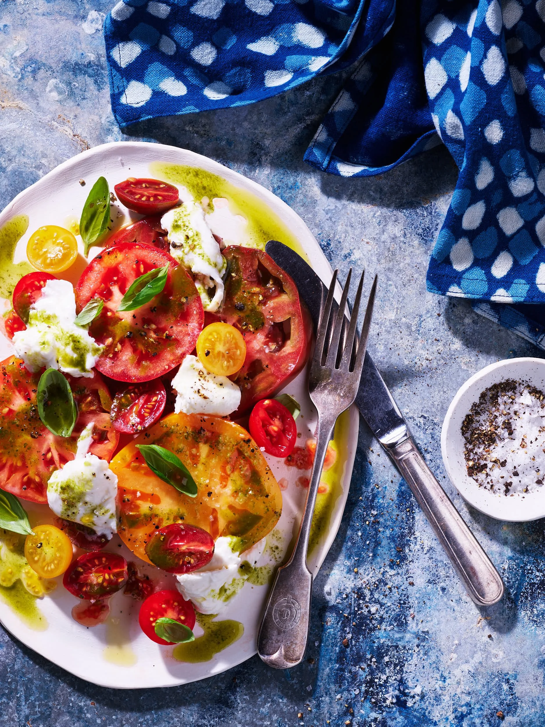 A plate of heirloom tomato salad with cherry tomatoes, fresh basil, mozzarella, and drizzled with olive oil and balsamic vinegar on a textured blue surface with a blue polka dot cloth and a small bowl of sea salt.