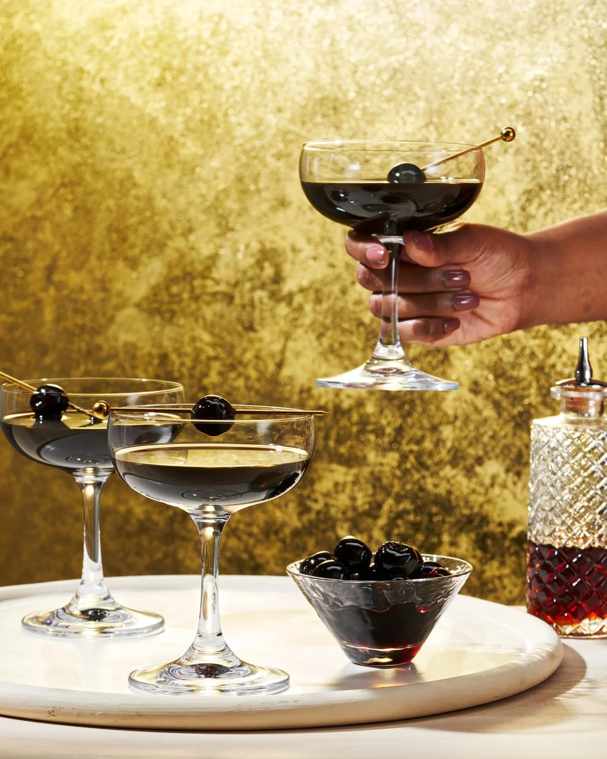 A hand holding a cocktail glass filled with a dark drink, garnished with a cherry on a toothpick. Three other similar glasses with dark drinks and cherries are on a white tray, along with a bowl of cherries and a vintage-style bottle of liquor agains