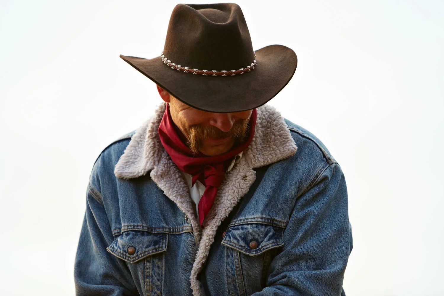 A man wearing a wide-brimmed cowboy hat, denim jacket with a fleece collar, and a red bandana around his neck, looking downward.