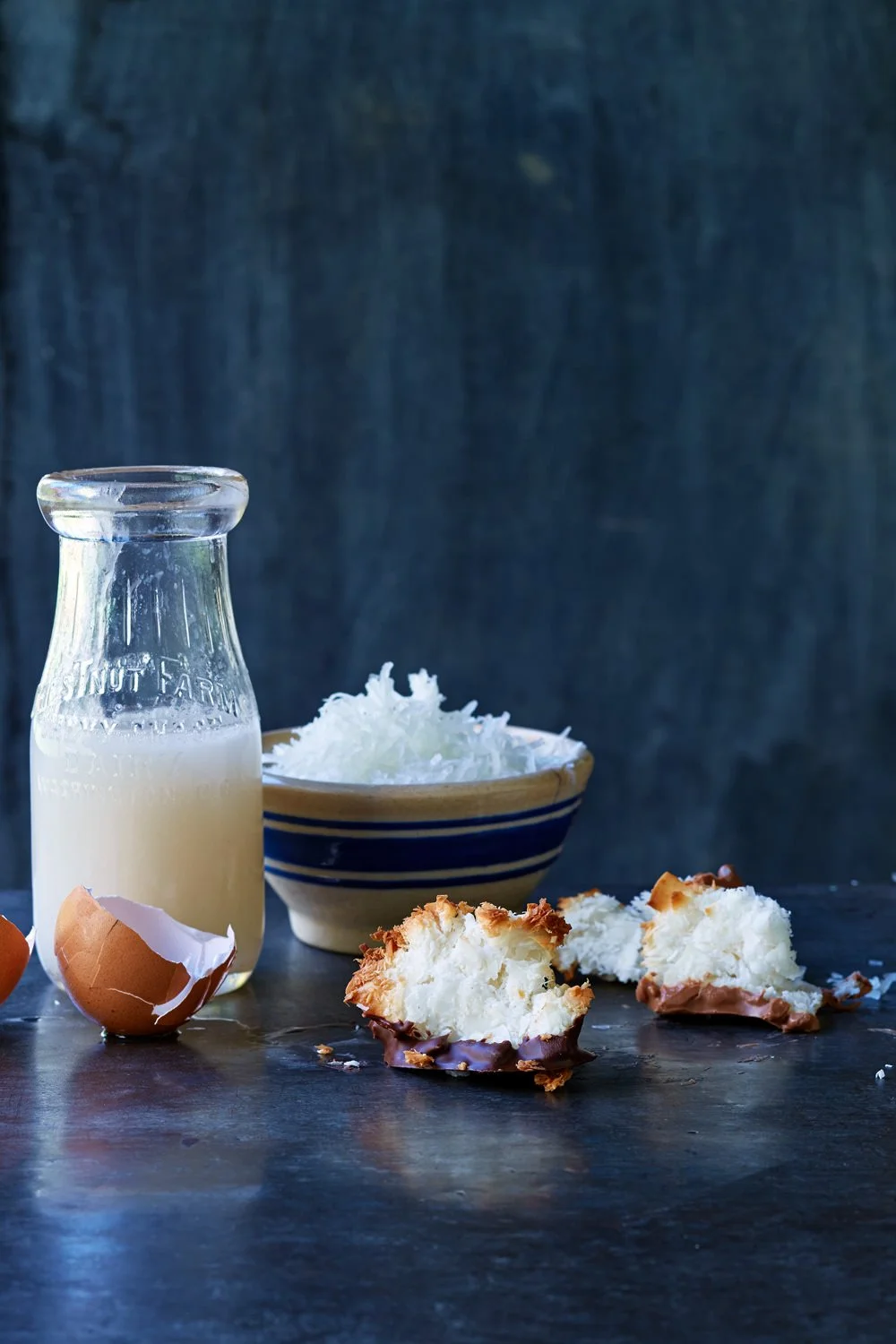 Broken eggshells, a small bottle of milk, a bowl of shredded coconut, and a coconut macaroon dessert on a dark surface.
