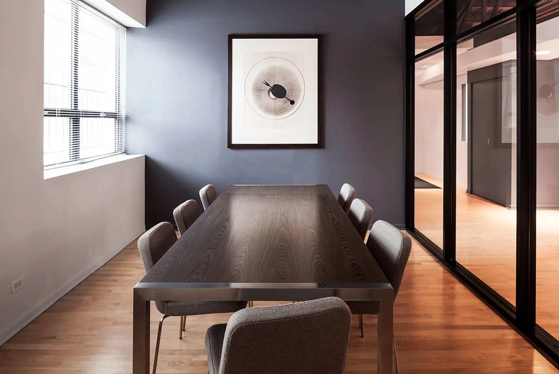 A modern conference room with a wooden table, grey chairs, a window with blinds, and a framed abstract art piece on a dark grey wall.