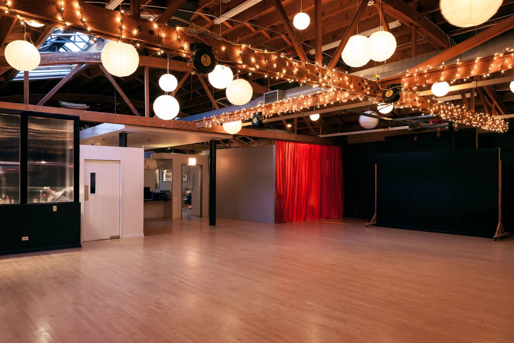 Empty event or dance hall with wooden floors, decorated with string lights and paper lanterns hanging from a wooden ceiling. There are red curtains and some partition walls.
