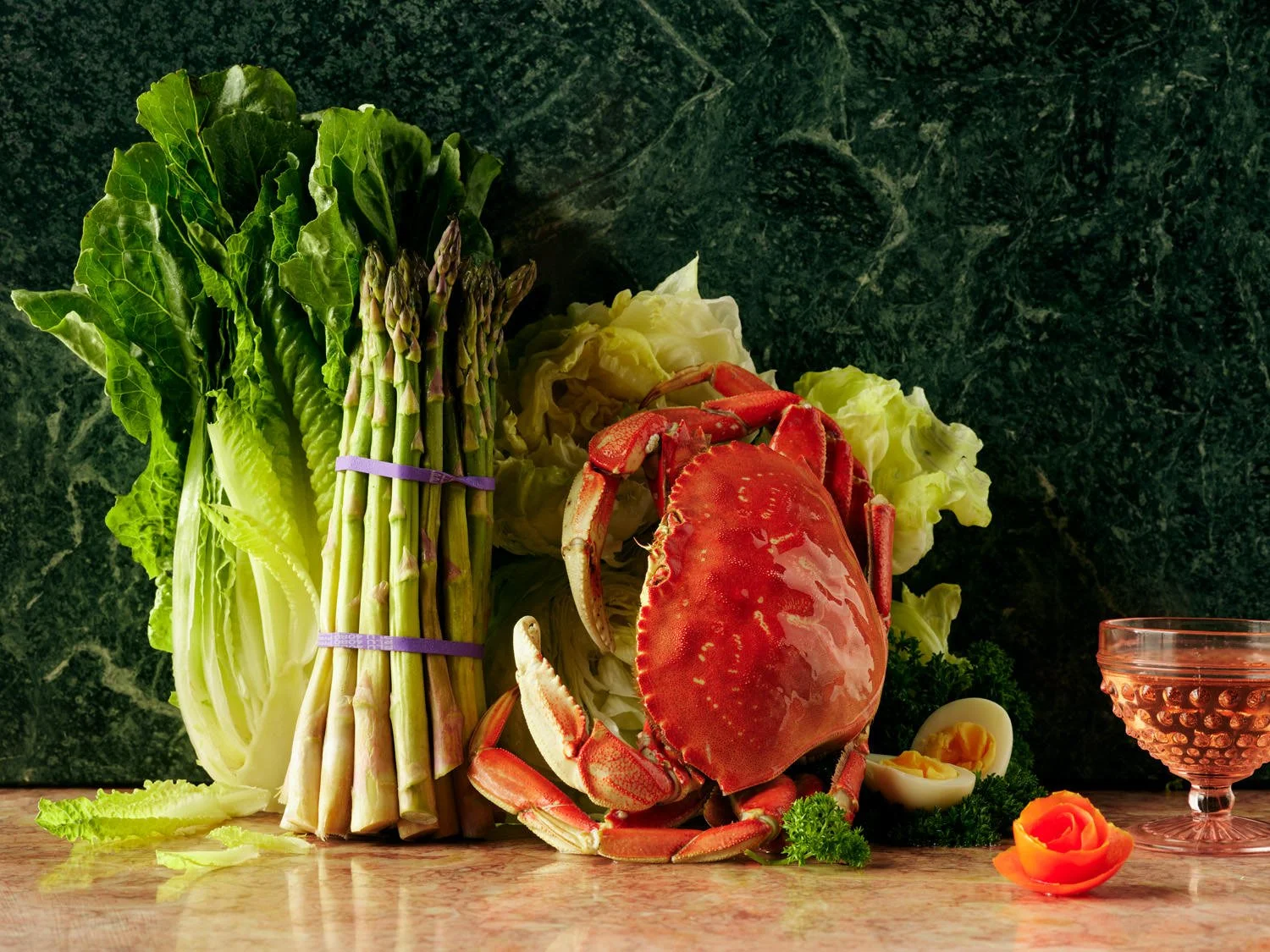 Fresh vegetables including lettuce, asparagus, and cabbage, with cooked crab, a soft-boiled egg, and a pink glass bowl on a marble surface.