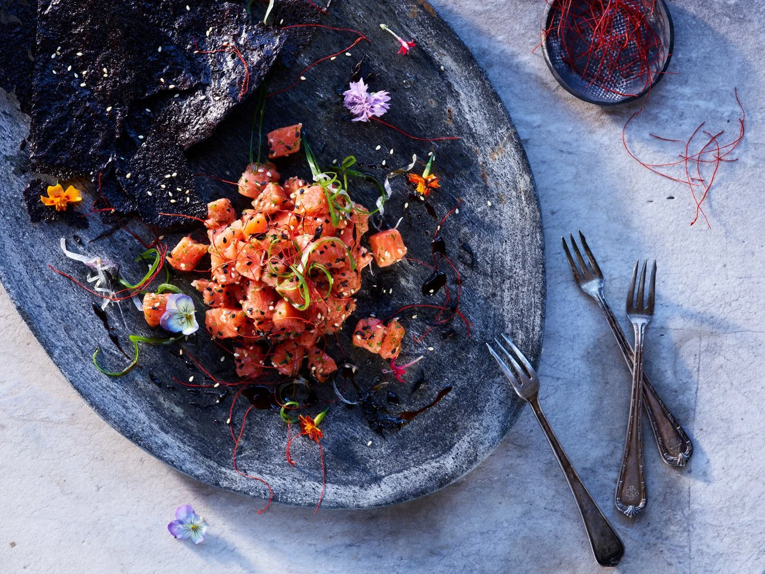 A plate of cubed raw salmon sashimi garnished with microgreens, sesame seeds, and edible flowers, served on a dark, round stone serving platter with black lace-like crisps nearby. Two vintage silver forks are placed to the right on a light textured s