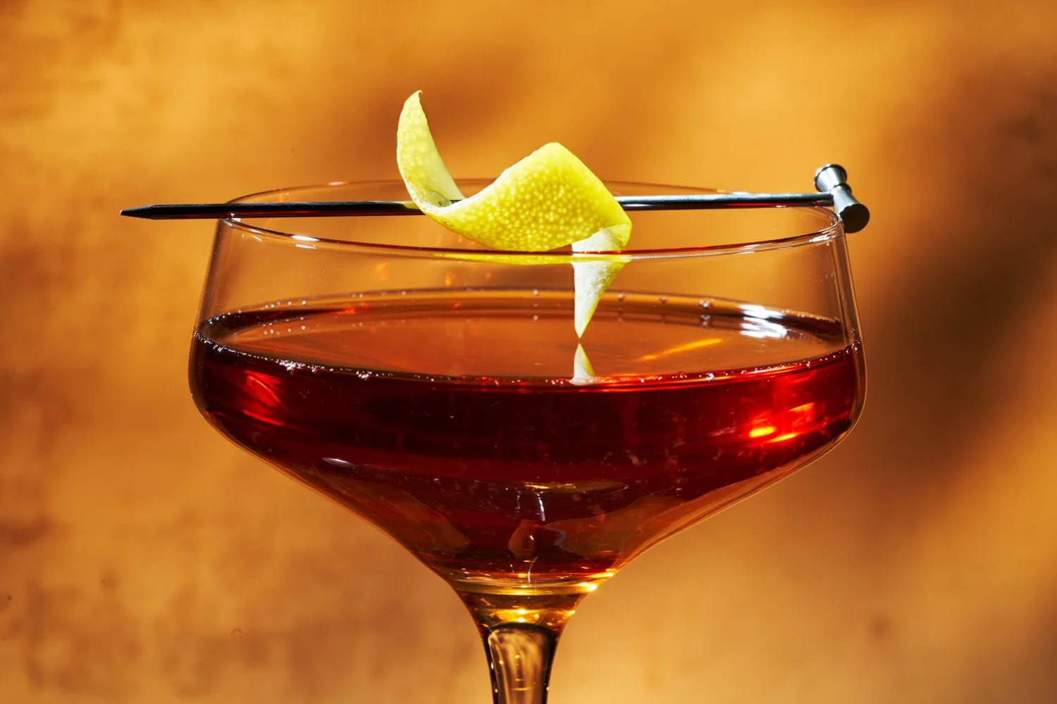 A cocktail glass filled with a dark red drink, garnished with a lemon twist, and a cocktail pick resting on the rim, against a warm, blurred background.