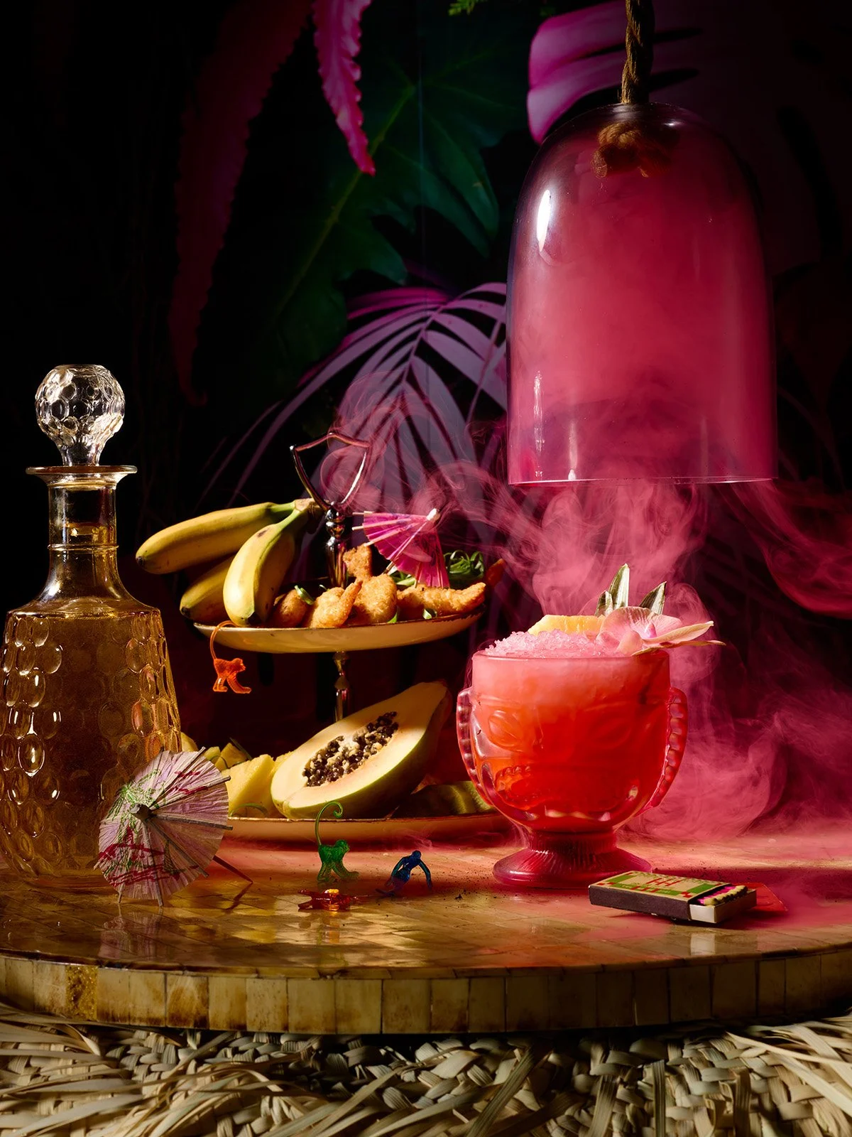 Colorful tropical-themed still life with fruit, drinks, and decorative items on a wooden table, with pink and green foliage in the background.