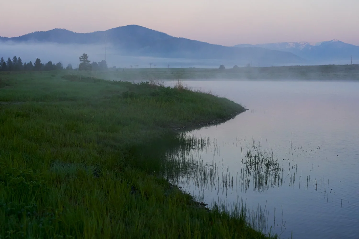 A peaceful lakeside scene with calm water, green grass along the shore, mist rising over the water, and mountains in the background, set during dawn or dusk.