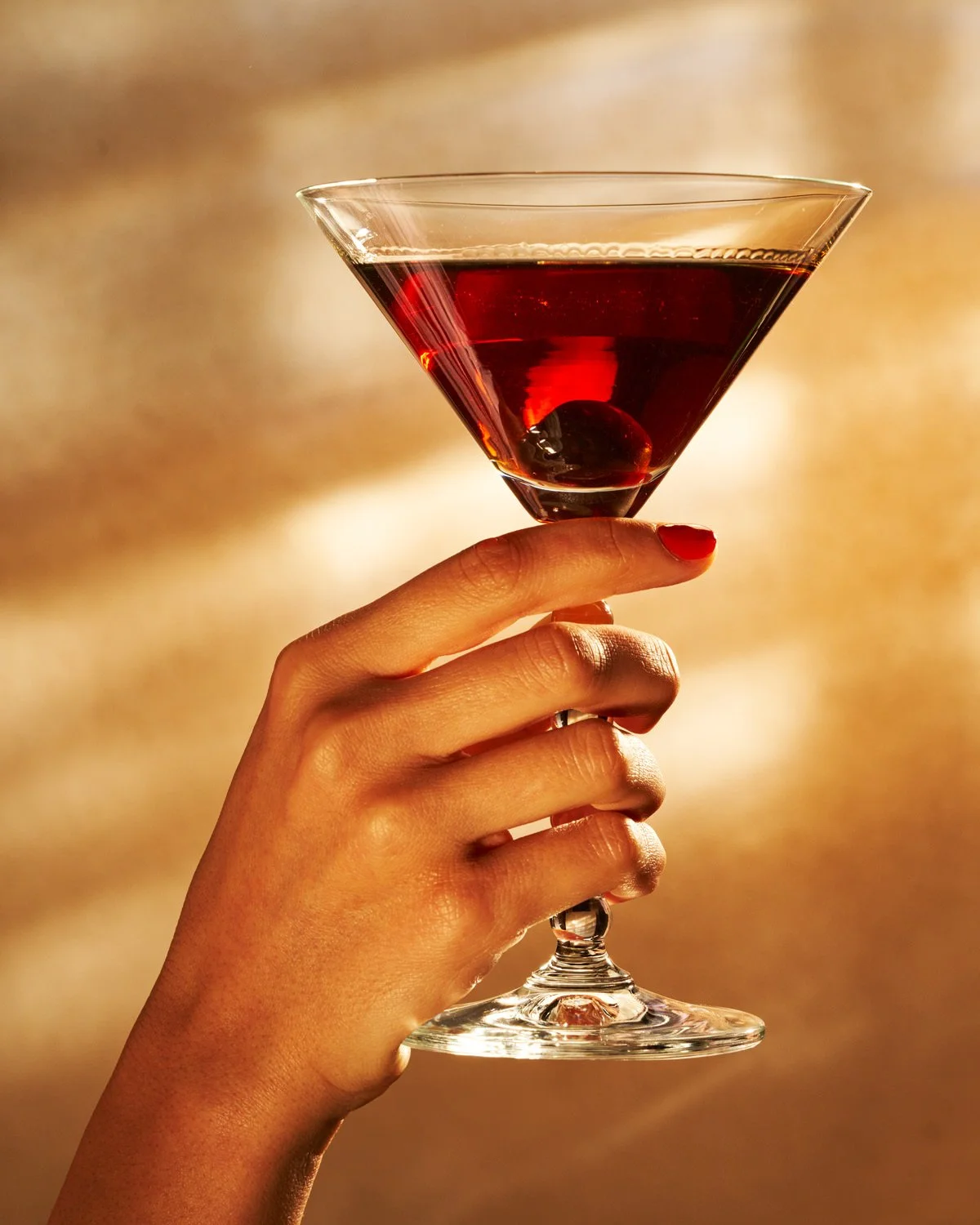 A hand with red nail polish holding a martini glass filled with a dark red cocktail and a cherry at the bottom, against a blurred warm background.