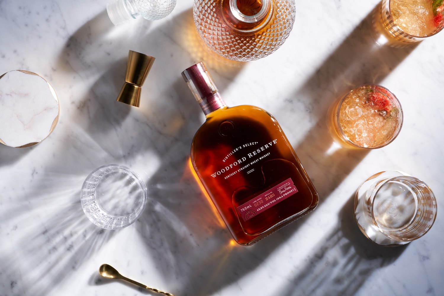 A bottle of Woodford Reserve bourbon whiskey surrounded by glasses of cocktails, a marble container, and a gold spoon on a white marble surface.