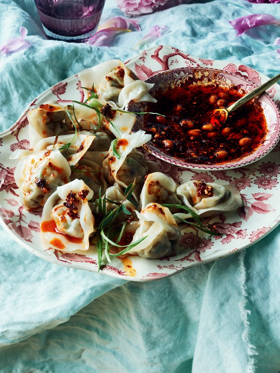 Plate of steamed dumplings garnished with green onions, served with spicy soy sauce in a decorative bowl, on a pastel blue tablecloth with a purple beverage in the background.