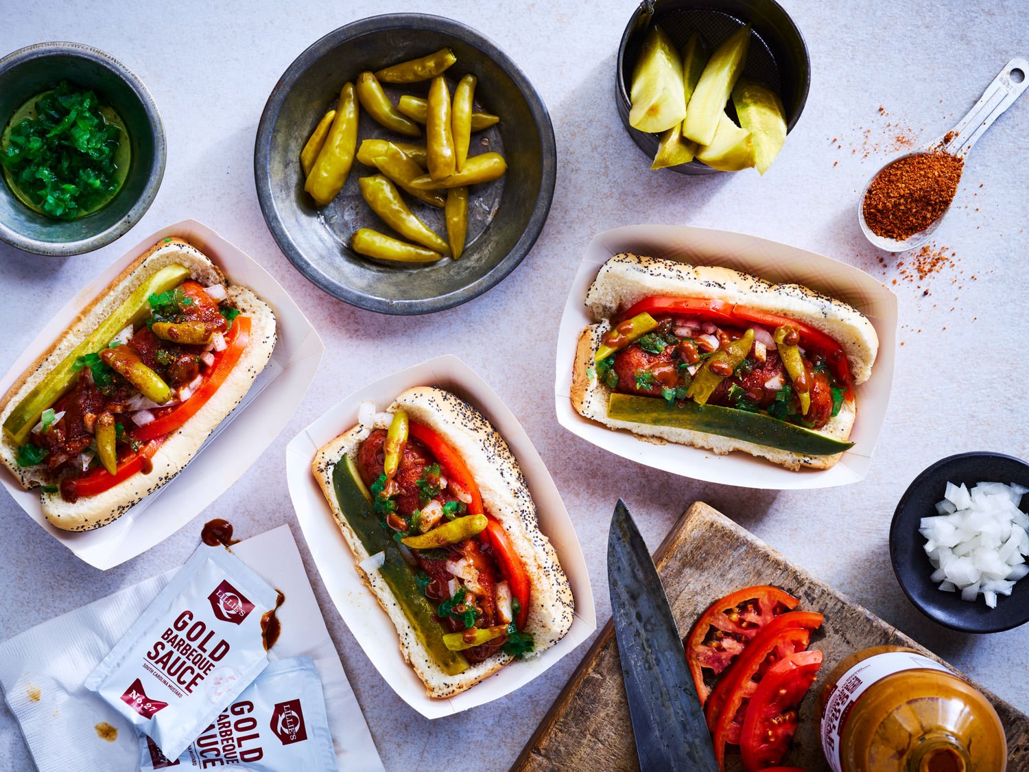 Three hot dogs with toppings including tomatoes, peppers, onions, and sauces, on white trays; bowls of sliced onions, pickles, chopped peppers, and slices of tomato; bottles of beer and packets of barbecue sauce; wooden cutting board and a knife.