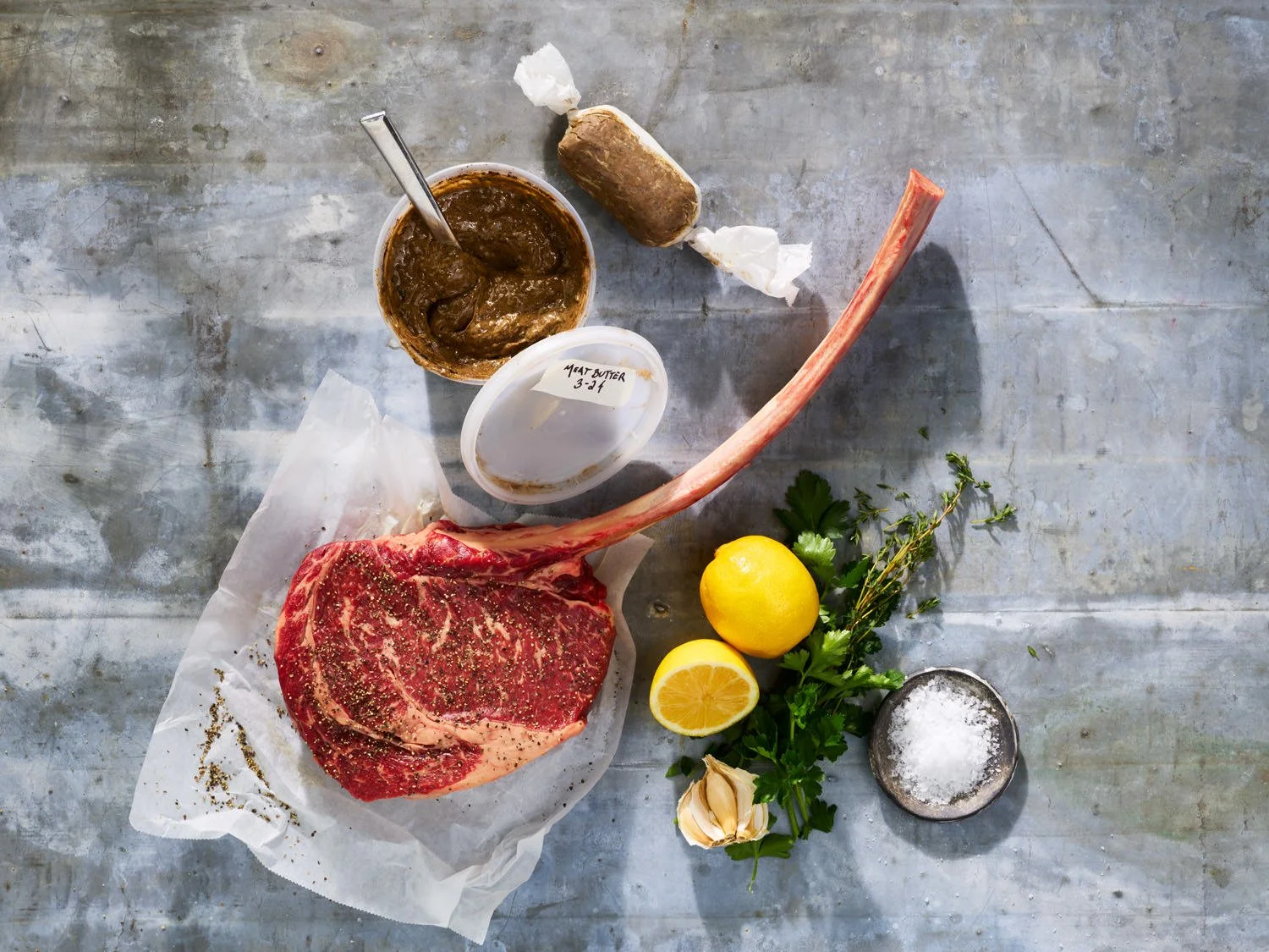Raw tomahawk steak with herbs, lemon, garlic, salt, and a cup of seasoned meat paste on a gray surface.
